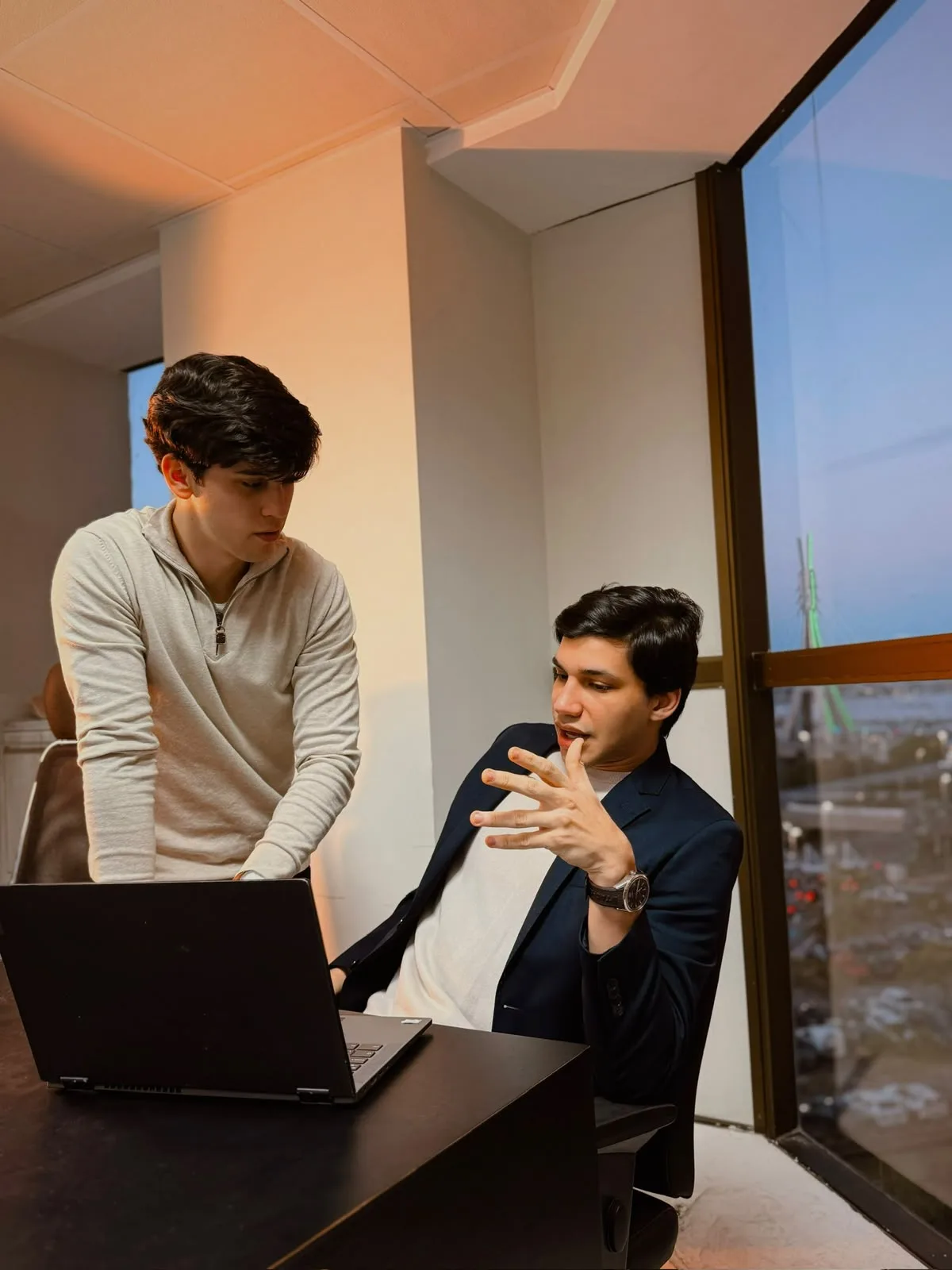 Os sócios da Axial conversando no escritório, um em pé e outro sentado diante do notebook, com vista da cidade pela janela